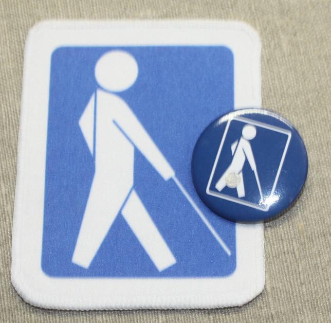 Näkövammaismerkit. Neliö kangasmerkki ja pyöeä rintanappi. Sinisellä pohjalla valkoinen kävelevä ihmishahmo ja keppi
