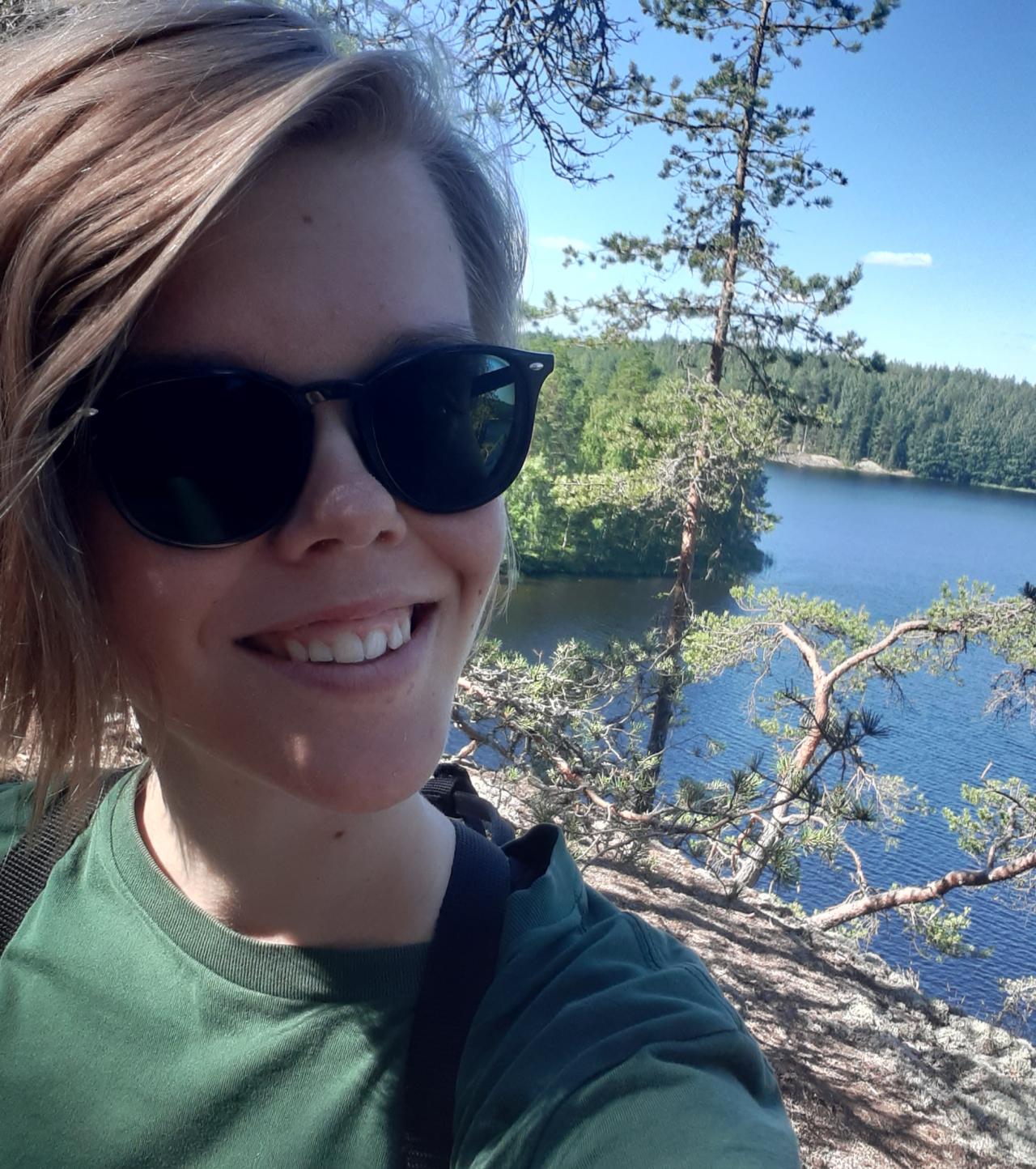 Anniina hymyilee, ja hänellä on aurinkolasit ja vihreä t-paita. Kuva on rajattu rintakehän korkeudelta. Taustalla näkyy kalliota, puita ja vettä.