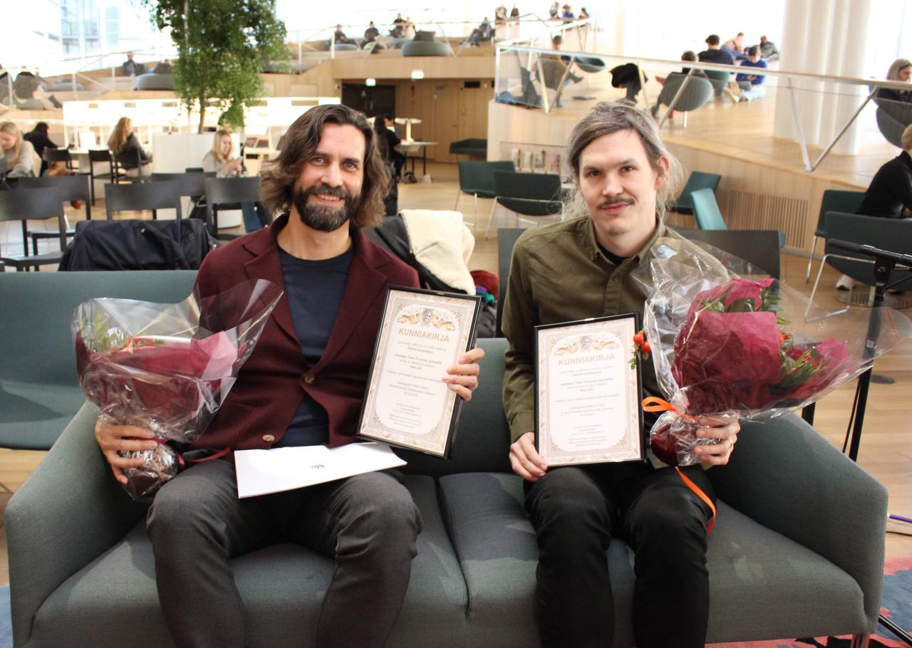 Kaksi miestä istuu sohvalla käsissään kunniakirjat ja kukkakimput, hymyilevät ja katsovat kameraan.