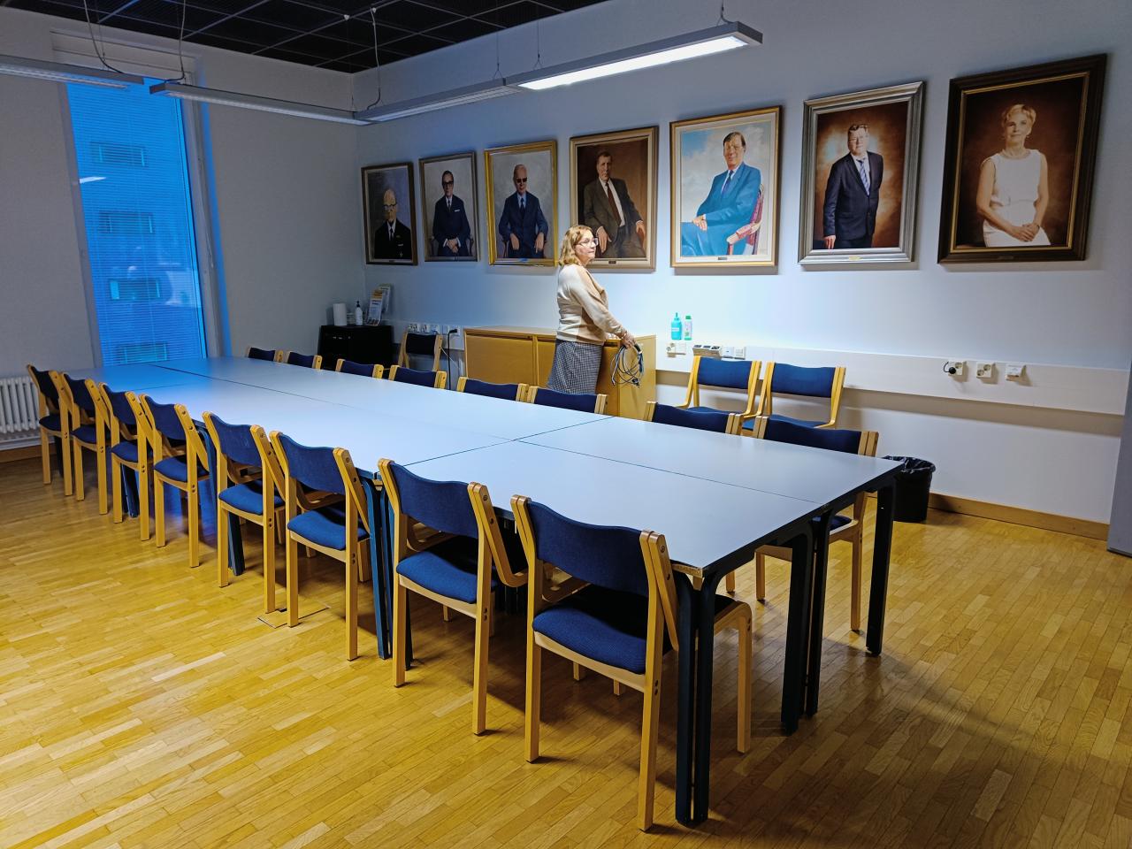 Kuvassa on kokoustila Mathildan kalustus. Vaaleat pöydät ja siniset tuolit. Seinällä on muotokuvatauluja hallituksen puheenjohtajista.
