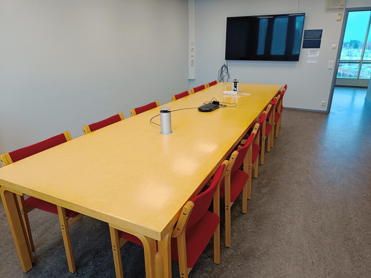 Kuvassa on kokoustila Einarin kalustus. Vaaleapuinen pöytä ja punaiset tuolit.