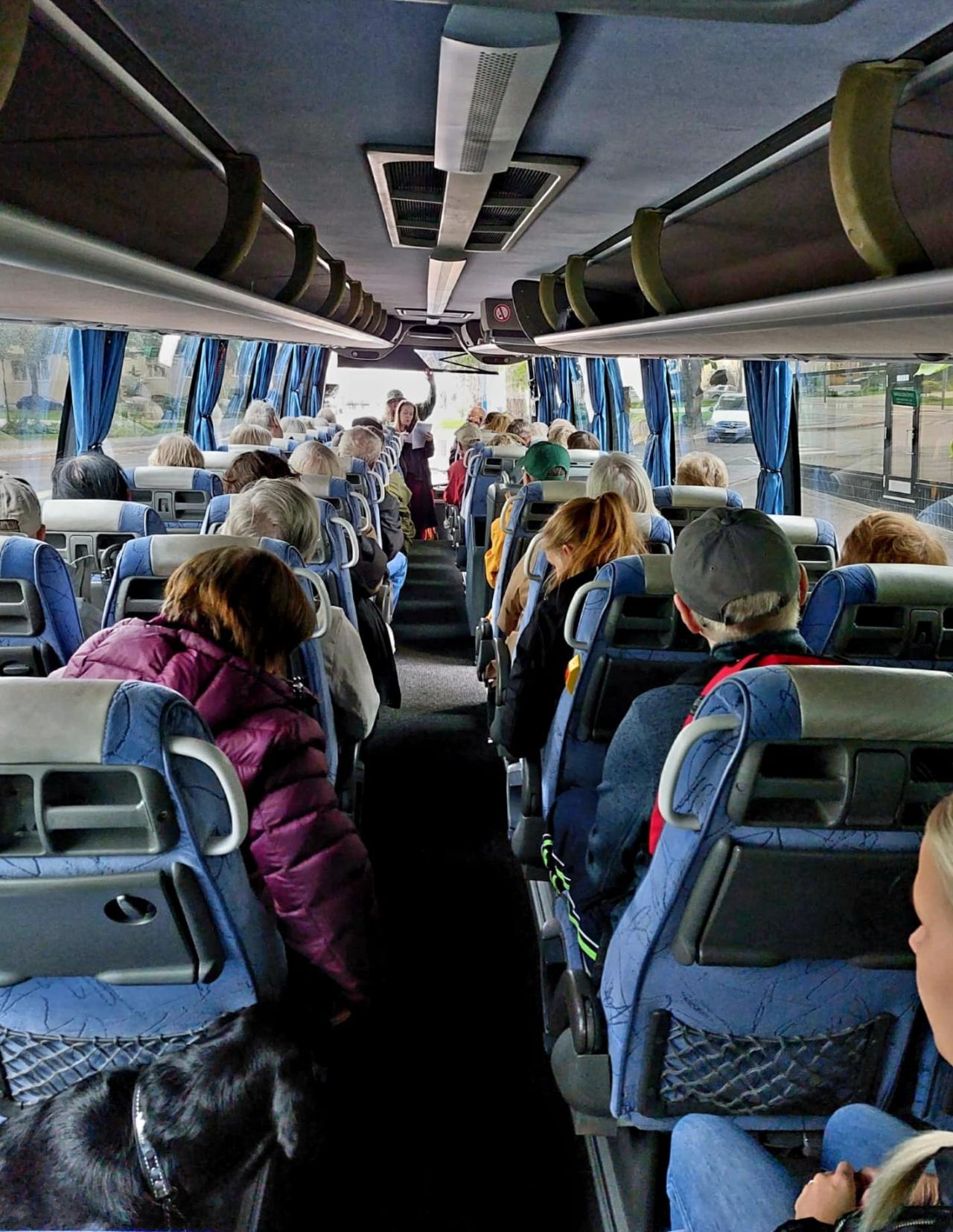Kuva otettu ison linja-auton takapenkiltä keskeltä. Molemmin puolin istuu ihmisiä kahdenhengen paikoilla, ja he katsovat kohti bussin etuosaa, jossa toimistotyöntekijä Tia toivottaa väen tervetulleeksi.