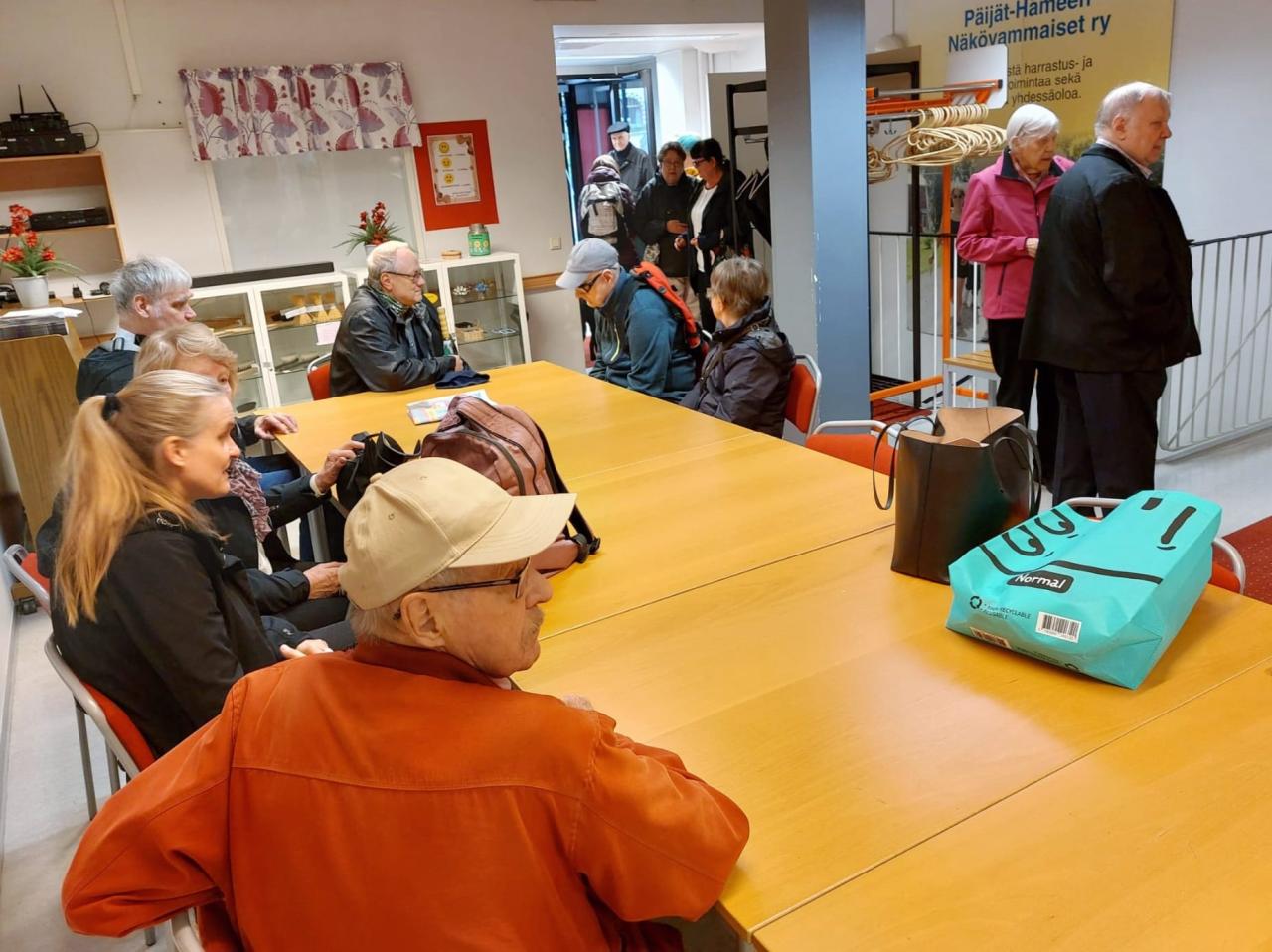 Pitkän puisen pöydän äärellä isossa salissa istuu seitsemän ihmistä tavaroineen. Taustalla näkyy toimintakeskuksen ovi, josta on juuri astunut sisään neljä henkilöä. Kaksi henkilöä riisuu ulkotakkeja oikeassa yläkulmassa.