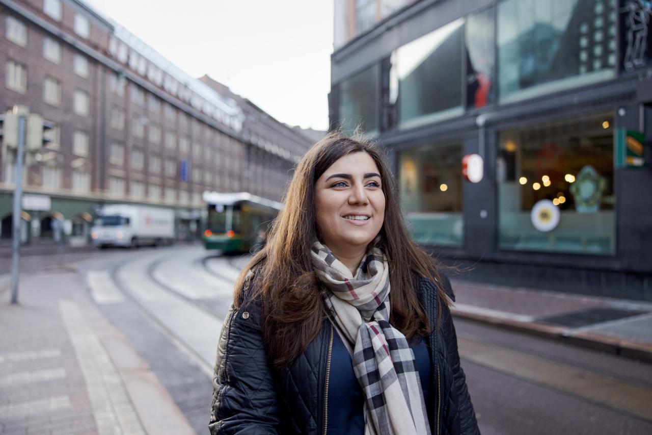 Nainen kävelee Helsingin keskustassa hymyillen. Etäällä taustalla näkyy pysäkillä oleva raitiovaunu ja raitiokiskot skeä muuta kaupunkiliikennettä.