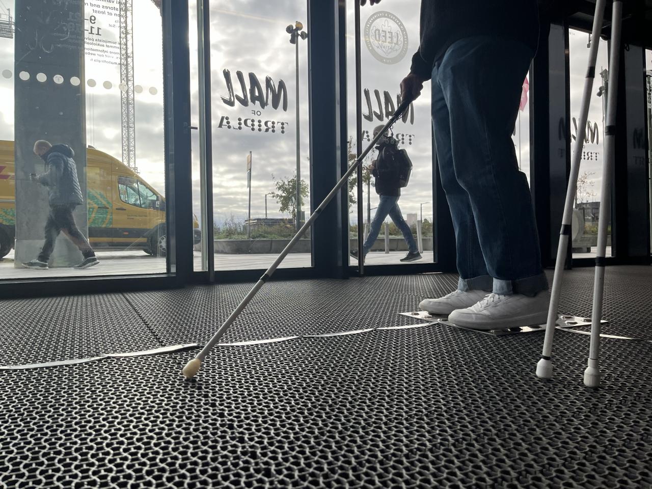 Lattiatasosta otettu lähikuva valkoista keppiä käyttävän henkilön jaloista Mall of Triplan tuulikaapissa.