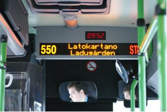 Bussin sisällä oleva iso pysäkkinäyttö, josta näkyy bussin numero ja seuraava pysäkki. Näytön yläpuolella on digitaalinen kello.