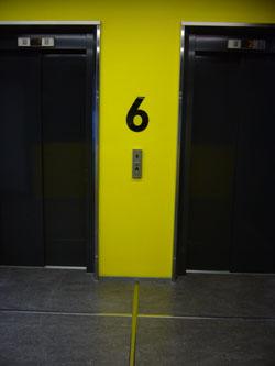 Kahden hissin välissä on keltaisella seinällä hyvin erottuva musta numero kuusi, joka kertoo kerroksen.