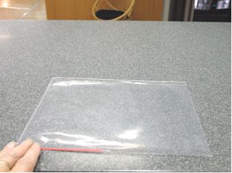 Harmaalla pöydällä on läpinäkyvä muovitasku. Vasemmassa alanurkassa näkyvät sormet, jotka pitävät kiinni muovitaskun nurkasta.