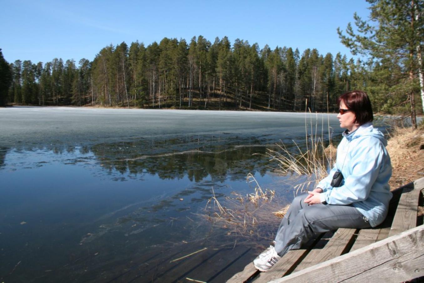 Kuvassa oikealla puolella nainen aurinkolaisessa istuu puisilla portailla järven rannalla. Ranta on sulaa, kauempana jäätä. Taustalla metsää ja sininen taivas.