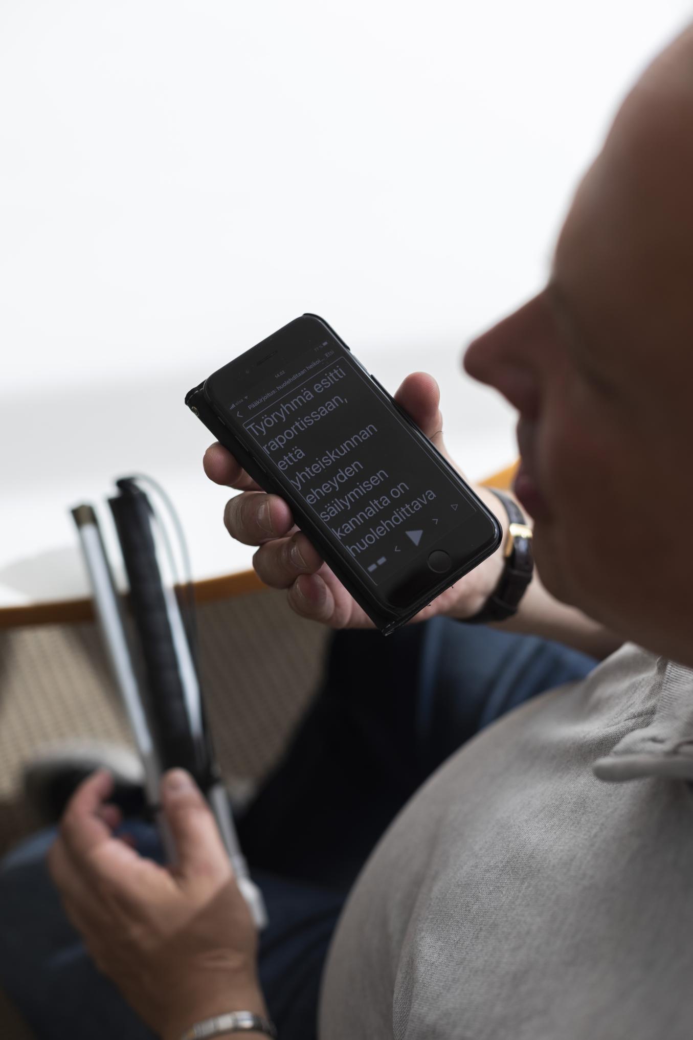 Lähikuva henkilöstö puhelin kädessä, ruudulla isotekstinen uutinen ja taustalla valkoinen keppi nojaamassa pöytään.