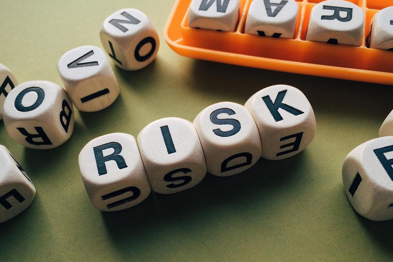 Puiset kirjainnopat muodostavat sanan 'RISK' vihreällä taustalla, ympärillä muita satunnaisia kirjaimia ja oranssi säilytyslokero.