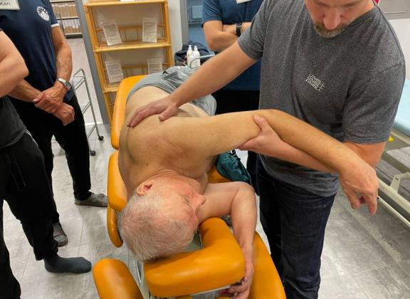 Miesasiakas makaa oikealla kyljellään ylävartalo paljaana. Kouluttaja Niclas Granfelt näyttää, miten hartiarenkaan aluetta käsitellään asiakkaan vasemman yläraajan avulla.