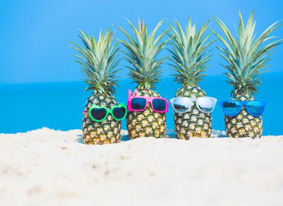 Neljä ananasta seisoo rantahietikolla aurinkolasit päässään. 
