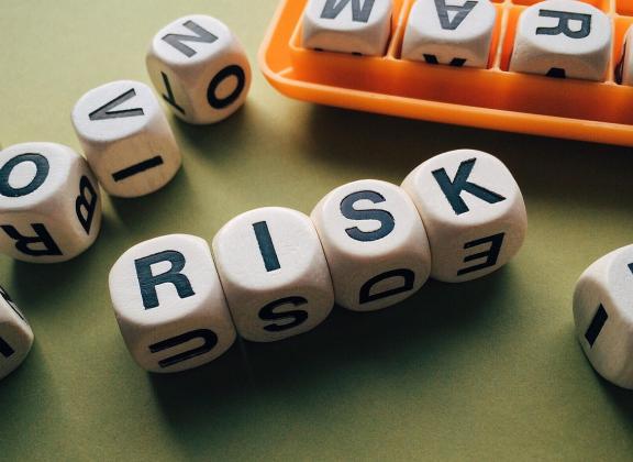 Puiset kirjainnopat muodostavat sanan 'RISK' vihreällä taustalla, ympärillä muita satunnaisia kirjaimia ja oranssi säilytyslokero.