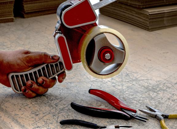 Lähikuva kädesta, joka pitelee teippikonetta kauniisti kuluneen puupöydän päällä.