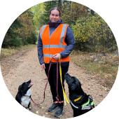 Henkilö ulkona metsäpolulla ja kaksi koiraa, joista toisella opaskoiranvaljaat.