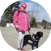 Henkilö pinkissä ulkoilutakissa ulkona valkoisen kepin ja opaskoiran kanssa.